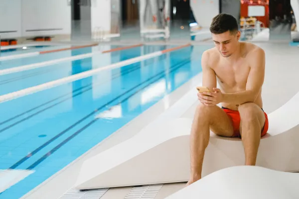 Nageur assis au bord d’une piscine intérieure consultant son téléphone.