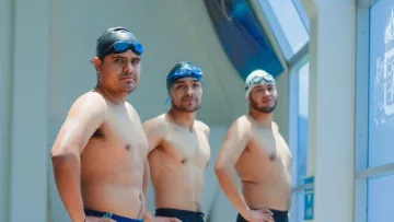 Three men stand in swimwear inside an indoor swimming pool area, ready for swimming activities