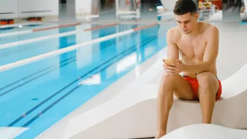 Nageur assis au bord d’une piscine intérieure consultant son téléphone.